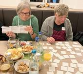 Auf dem Foto sind zwei sitzende Frauen an einem Tisch zu sehen. Vor stehen stehen Knabbereien, Getränke und Memorykarten liegen auf dem Tisch. Eine der Frauen liest gerade eine Spielanleitung.